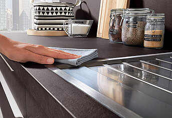 Eine Person wischt eine moderne Küchenarbeitsplatte neben einem Glaskeramikkochfeld, im Hintergrund sind Aufbewahrungsgläser und eine Kaffeetasse zu sehen.