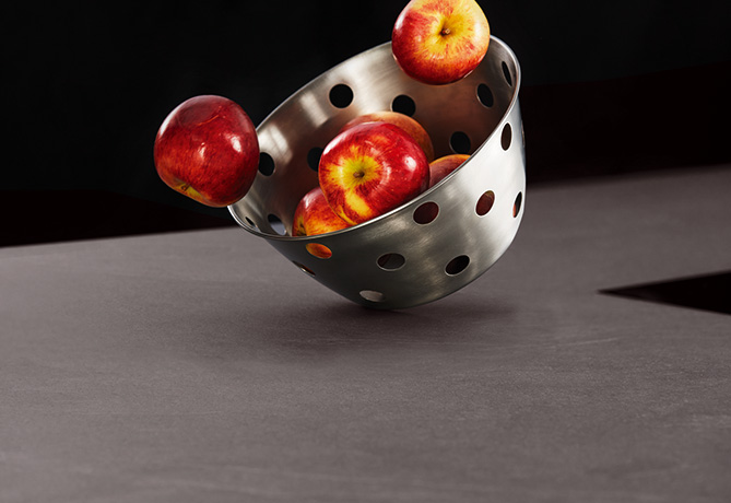 Ein Edelstahlsieb kippt um und lässt reife rote Äpfel auf eine dunkle kontrastierende Oberfläche fallen, die eine Mischung aus kulinarischer Nützlichkeit und frischen Produkten zeigt.